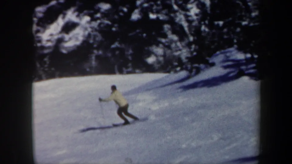 In Vermont 1962 home-movie footage, a distant skier in a light jacket and dark pants moves across a snowy slope bordered by evergreens.