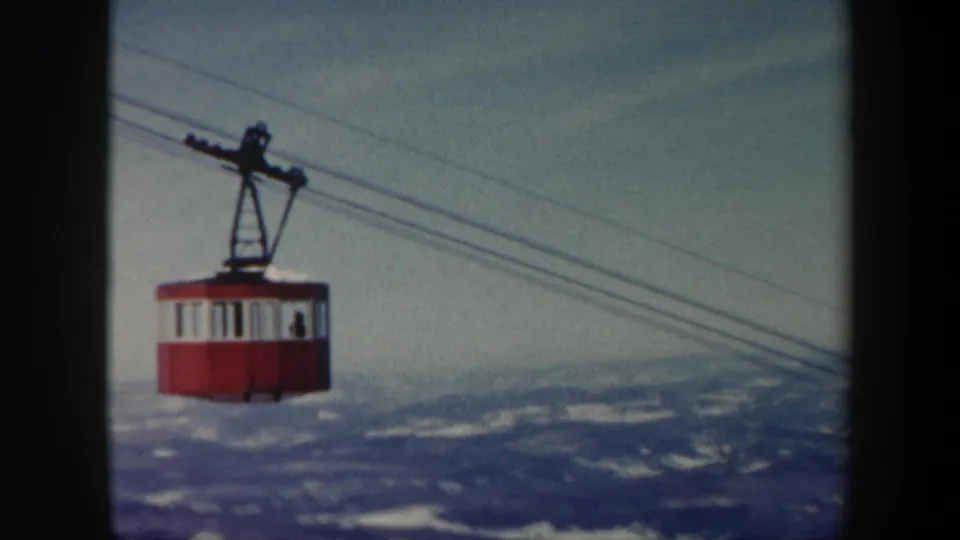 1962 Vermont 8mm view of a red cable car riding diagonal cables over snow-streaked hills.