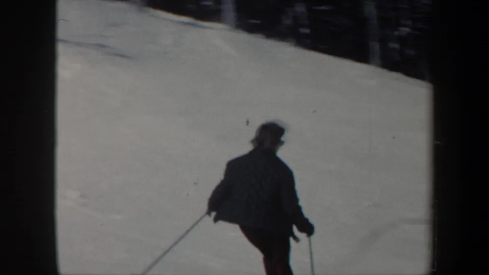 The skier is filmed from behind on a wide snowy Vermont slope, with poles angled outward and forest at the edge of frame.
