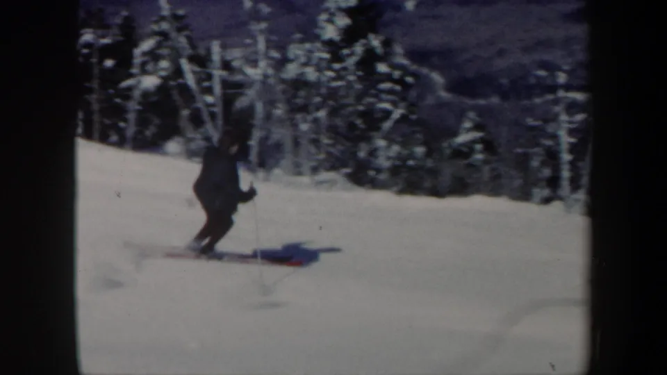 A dark-clothed skier turns on a snowy Vermont slope, with snow-covered evergreens and distant mountain terrain in the background.