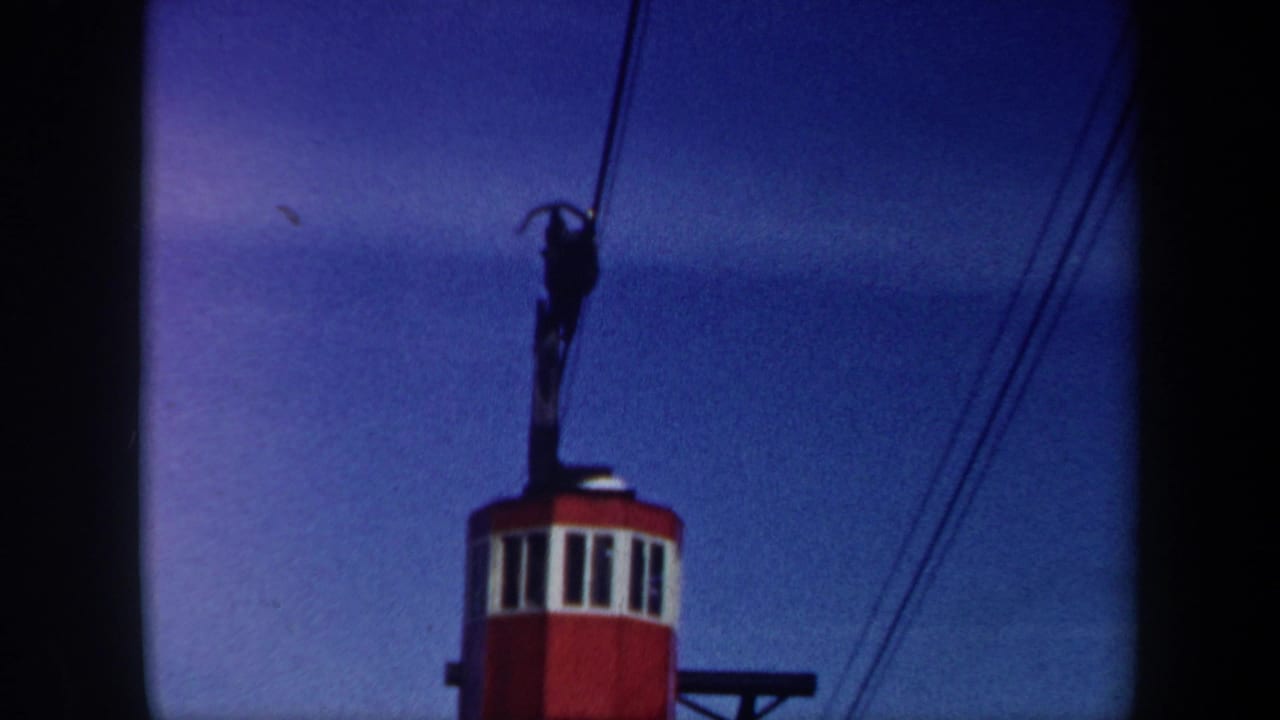 1962 Vermont 8mm frame showing a red and white cable-car cabin rising beneath dark overhead cables against a blue sky.