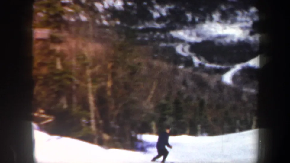 A lone skier in dark clothing moves across a snowy Vermont slope, with forested mountain terrain in the background.