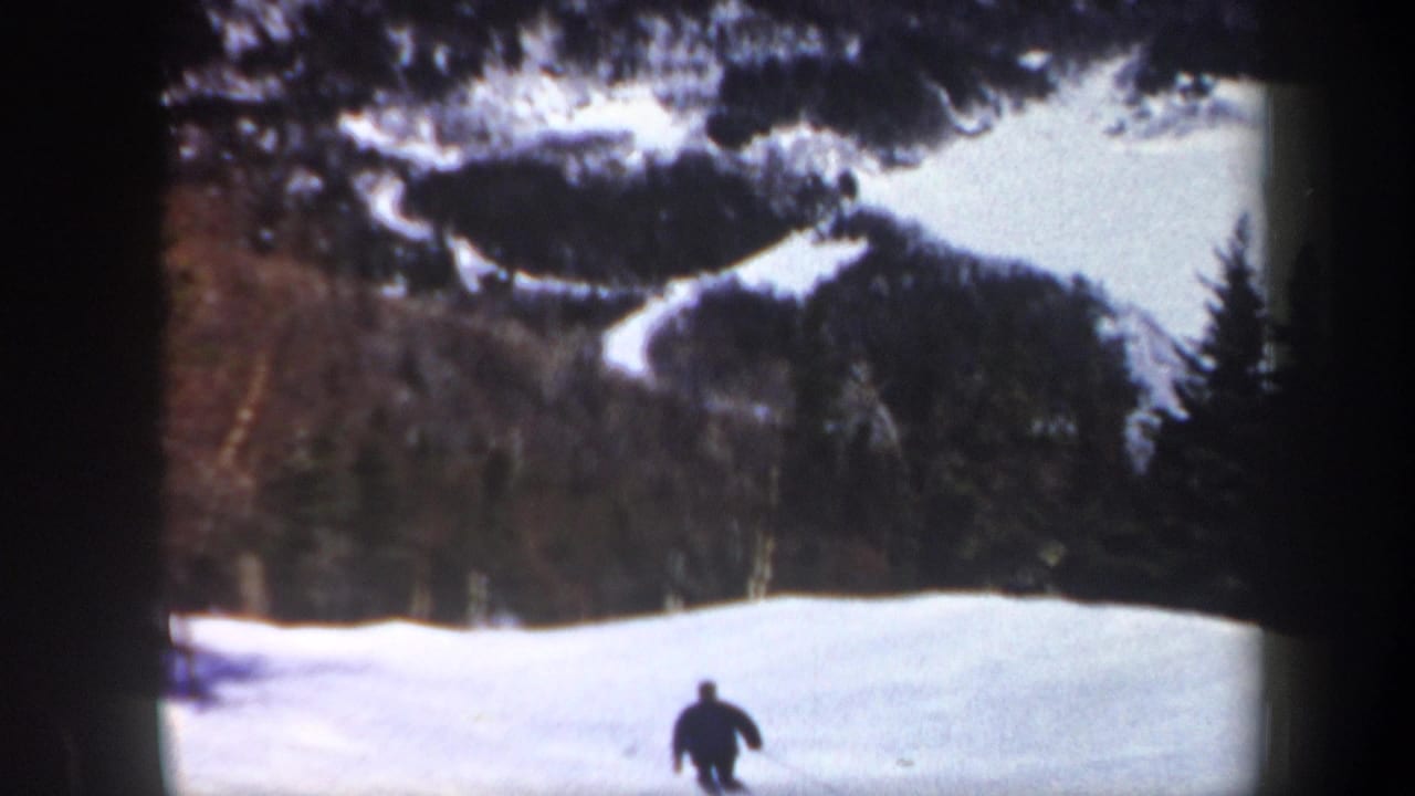 1962 Vermont 8mm frame with a lone skier moving downhill across a snowfield bordered by dark trees and wooded mountain slopes.