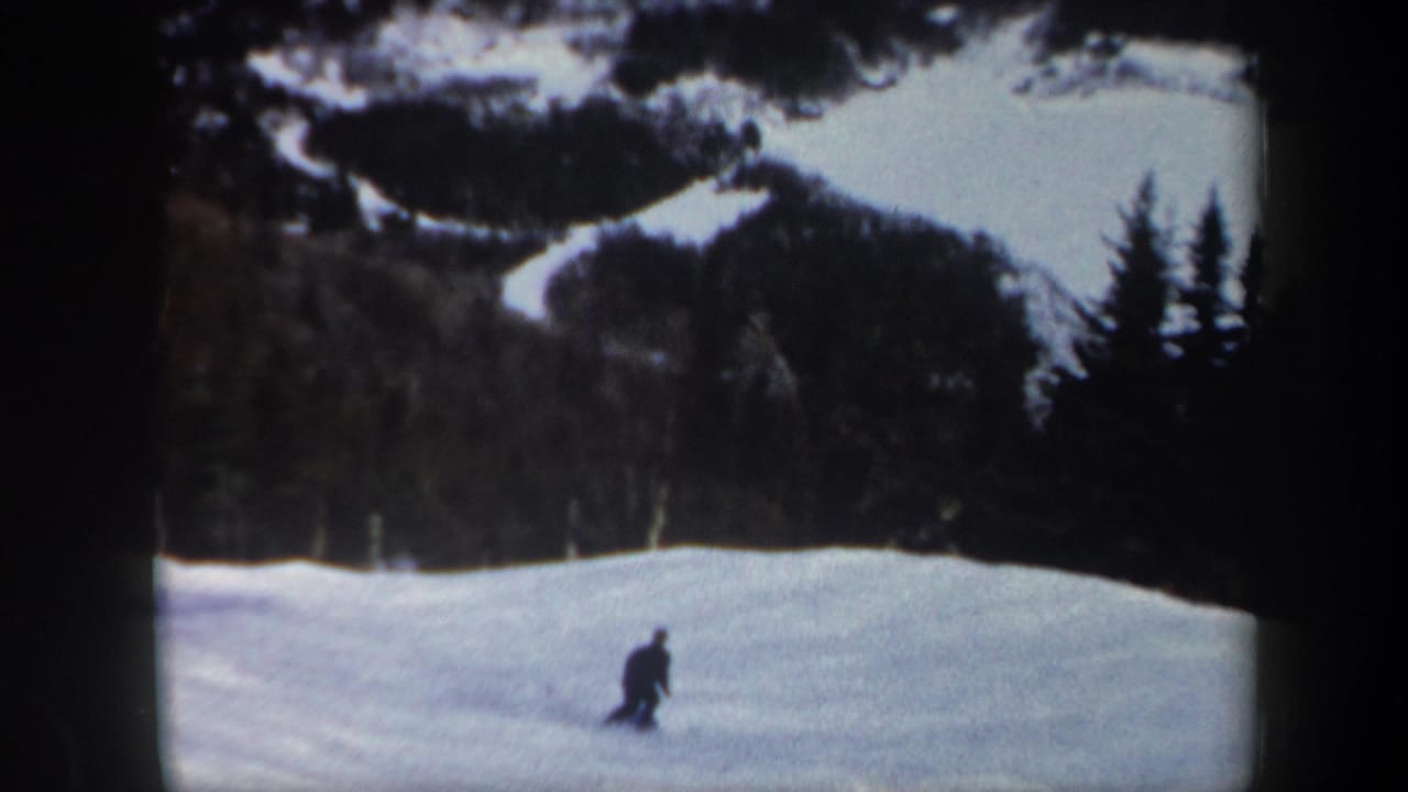 1962 Vermont home-movie frame showing a small, dark-clad skier carving across a snowy slope near a stand of evergreens.