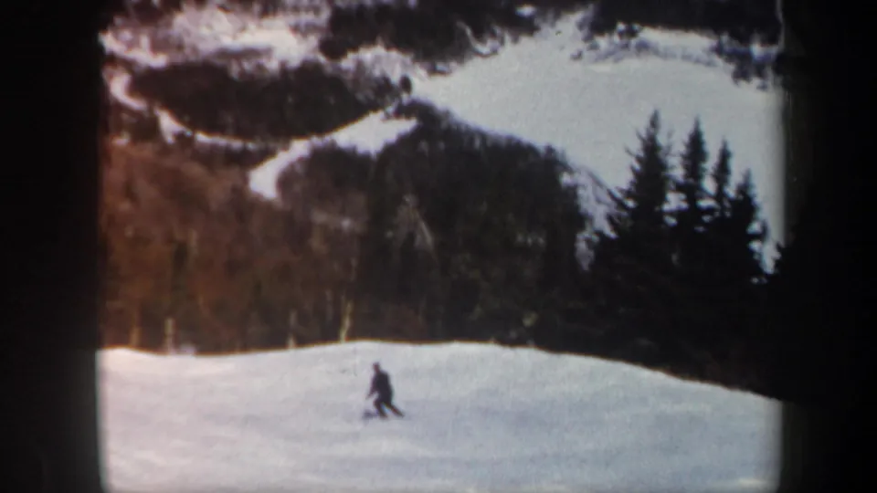 1962 Vermont 8mm still with the skier lower on the slope, moving past a forested backdrop and dark evergreen trees.