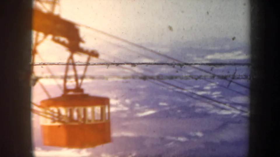 1962 Vermont frame with a red gondola cabin crossing cables above a hazy snow-covered mountain landscape.