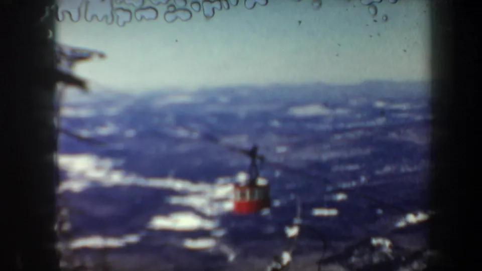 Wide 1962 Vermont 8mm view of a red gondola moving away across cables above a snow-spotted mountain valley.