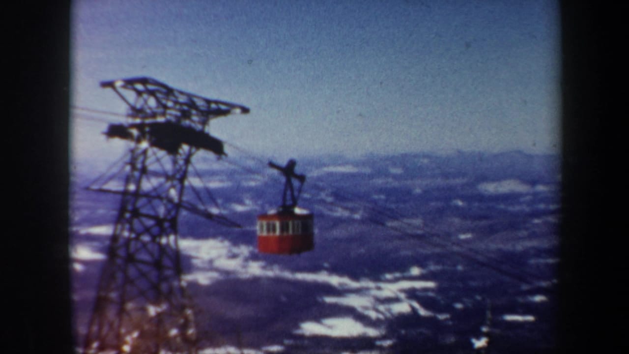 1962 Vermont home-movie frame of a red gondola moving past a dark steel lift tower over distant winter hills.