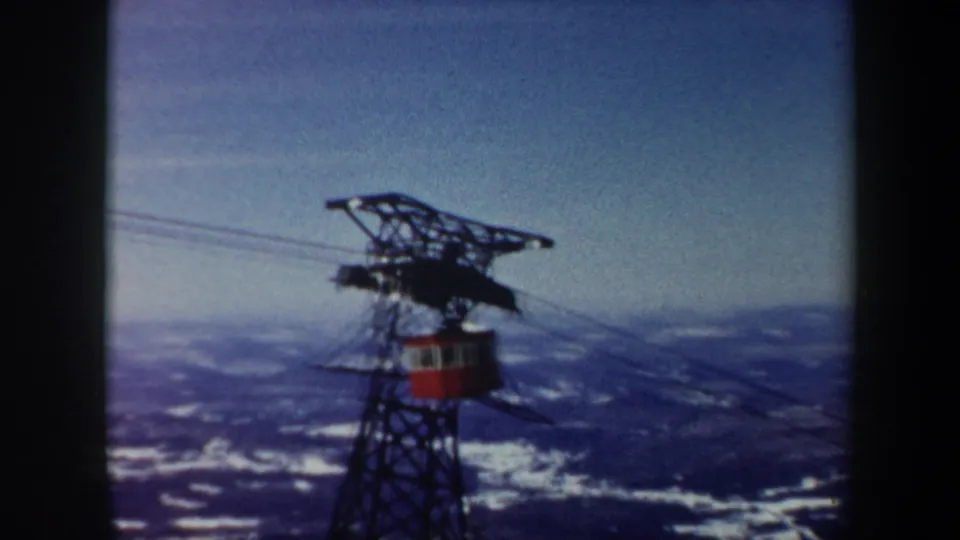 1962 Vermont 8mm frame showing the red cable car aligned near the lift tower, with hazy snowy mountains beyond.