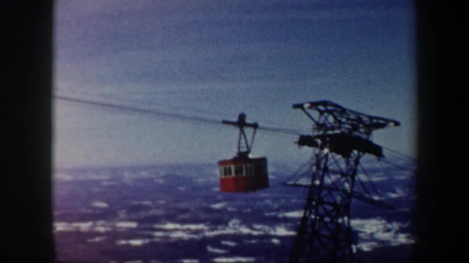 1962 Vermont home-movie frame of a red cable car moving away from a steel tower across a winter mountain backdrop.