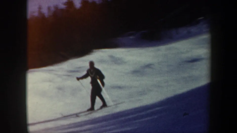 A skier in dark winter clothing uses poles on a snowy Vermont slope, with trees and strong shadow areas visible in the 1962 home-movie frame.