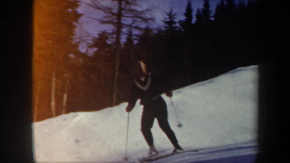 A distant skier with poles moves along a bright snowy slope bordered by dark conifer trees in Vermont 1962 8mm footage.