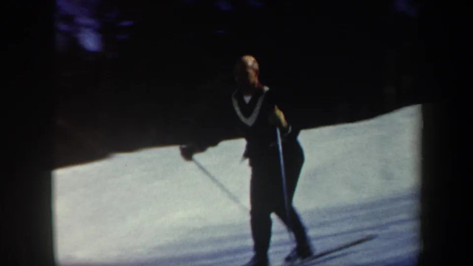 Adult skier in 1962 Vermont footage stands or moves on a sunlit snowy slope with dark evergreen trees in the background.