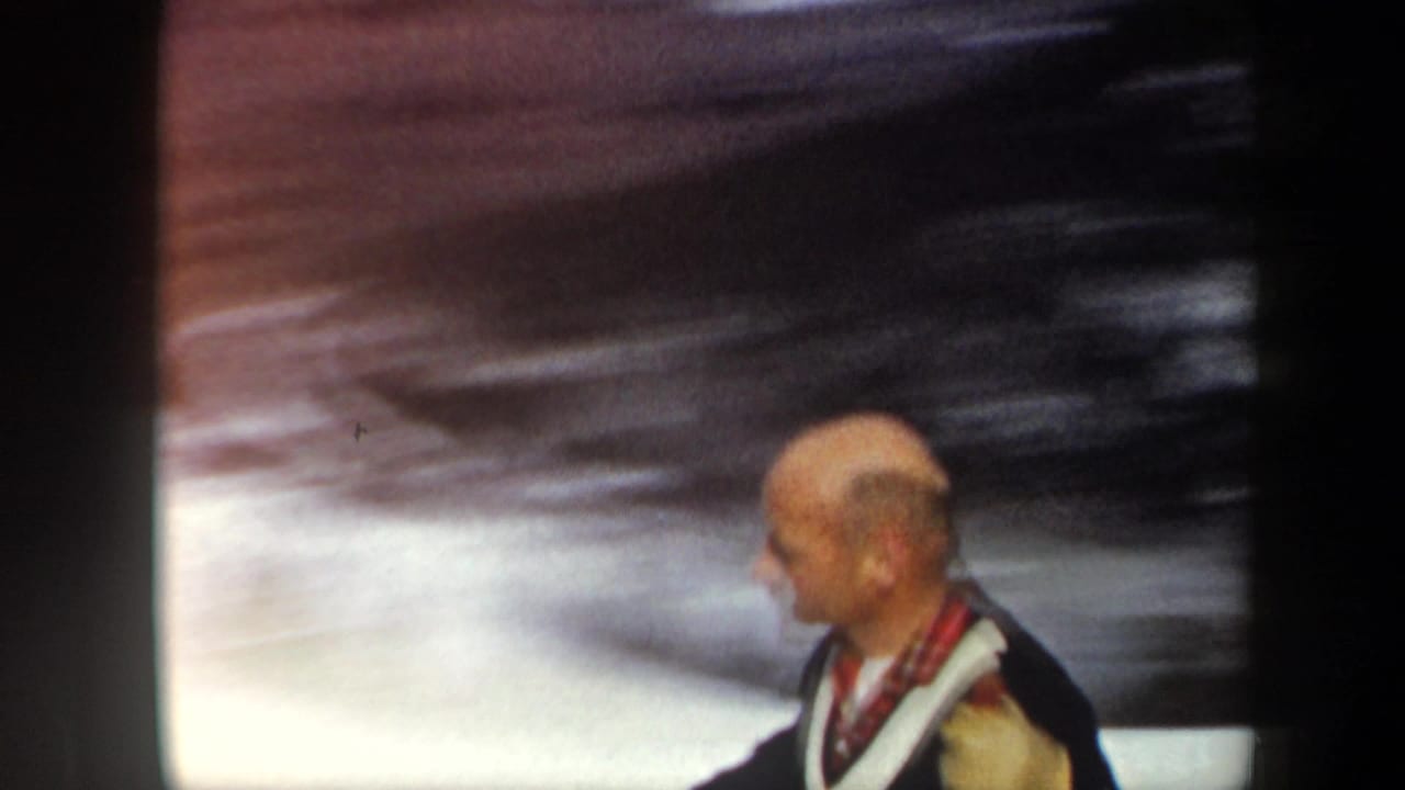 Close 1962 Vermont home-movie frame of a bald adult skier in a dark sweater with white trim and a plaid scarf.