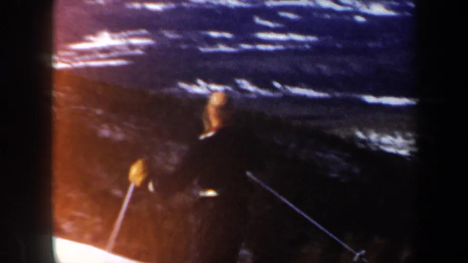 Adult skier in vintage 8mm Vermont footage uses poles while descending a snow slope near evergreen trees.