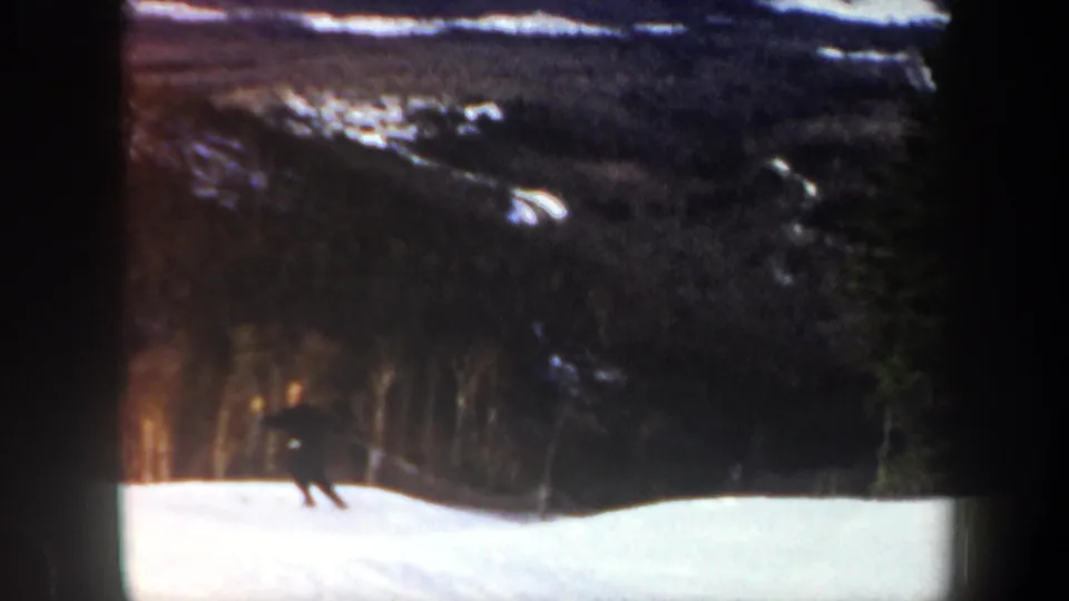 Grainy 8mm frame from Vermont in 1962 showing a skier low on a snowy slope with forest above.