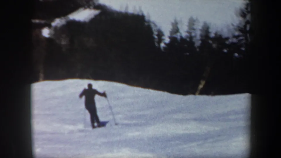 A distant skier crosses a bright snowfield below a dark wooded ridge in 1962 Vermont home-movie footage.