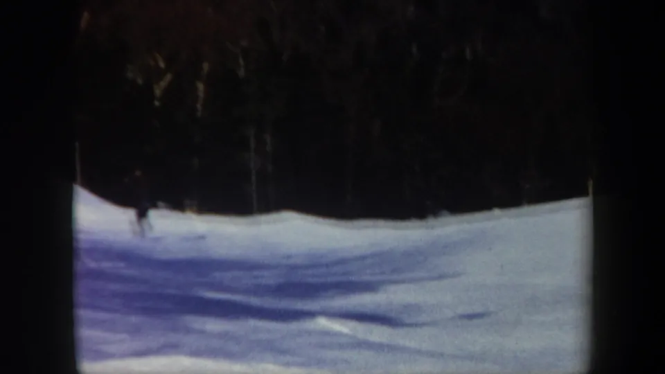 Home-movie frame from Vermont showing a small skier on a shaded snowfield below a dense, dark winter treeline.