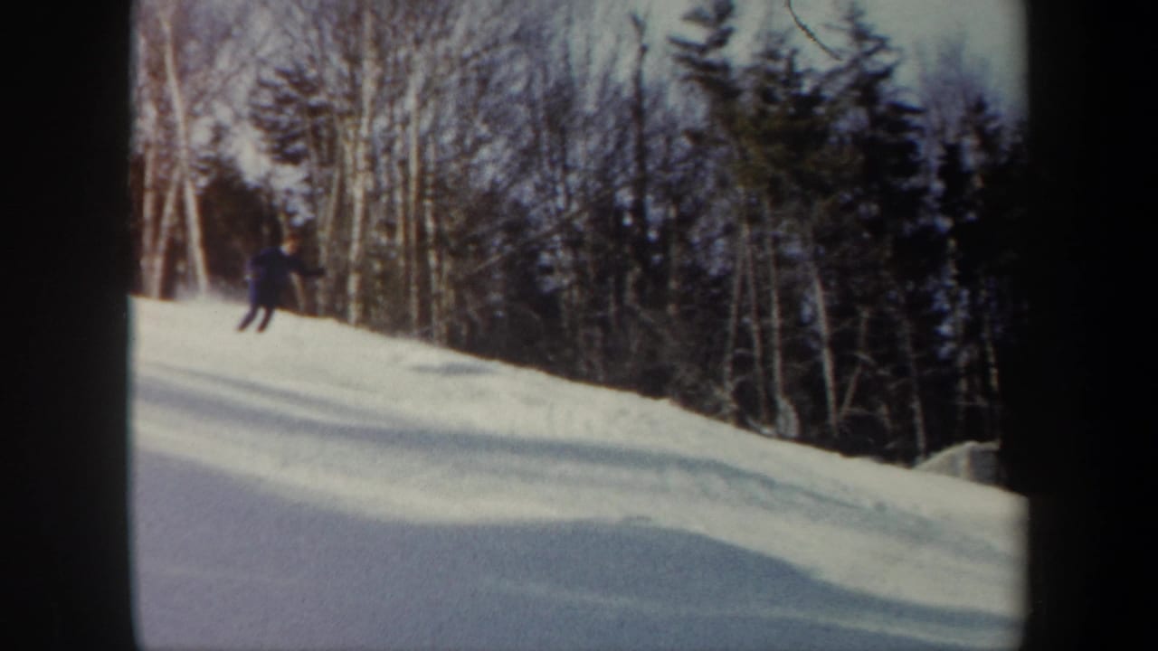 1962 Vermont home-movie frame with a skier in dark clothing turning on a snowy slope against birch and evergreen forest.