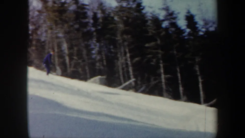 1962 Vermont 8mm frame showing a skier near the left edge of a snow-covered slope below dense evergreens and bare trees.