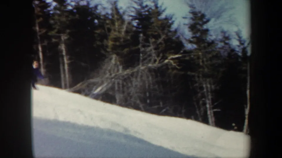 1962 Vermont home-movie frame with the skier partly obscured near a snow ridge and a backdrop of evergreens and bare branches.