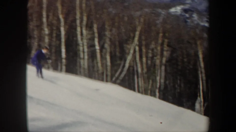 1962 Vermont 8mm frame of a skier in purple moving downhill across snow with a winter forest in the background.