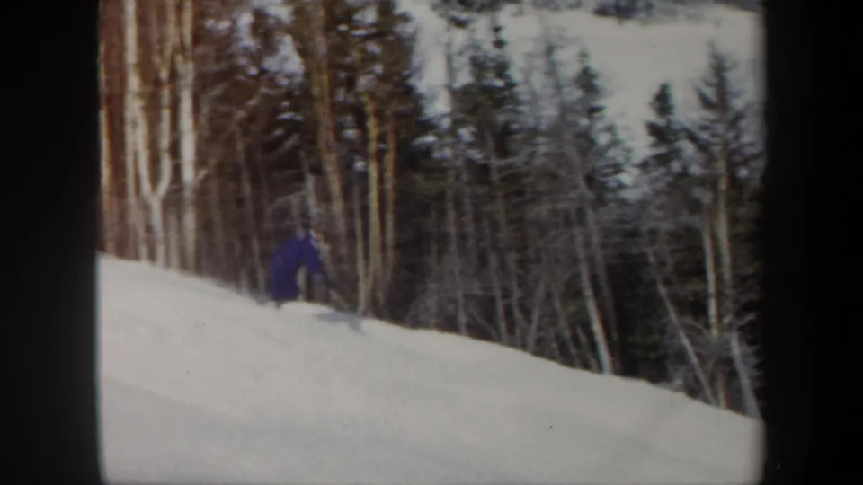 A distant skier in blue moves across a snowy wooded slope in archival 8mm footage.