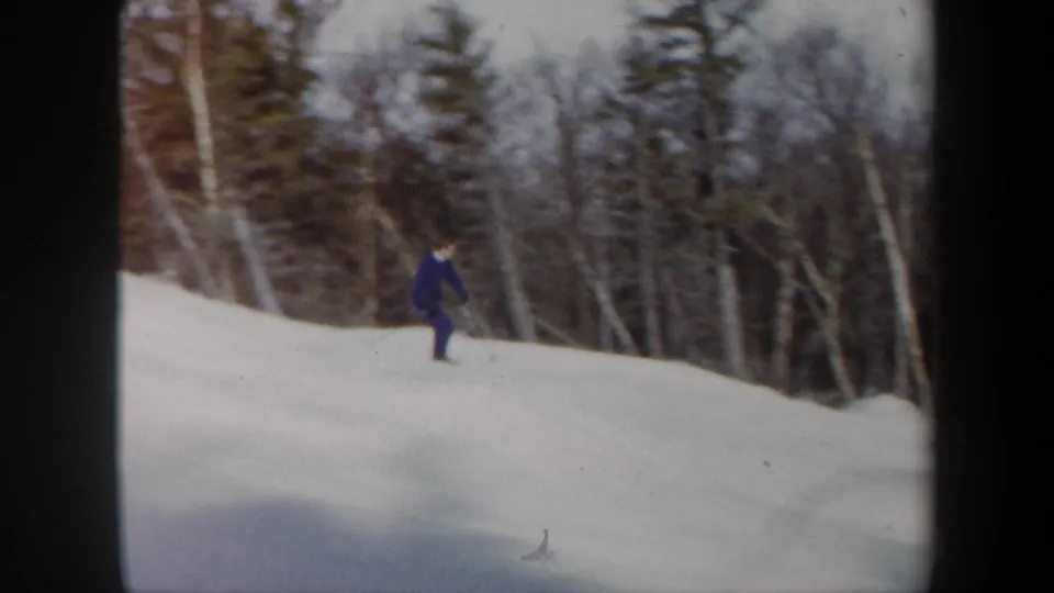 The skier continues across the snowy hill, framed by bare trees and evergreens in soft 8mm color.