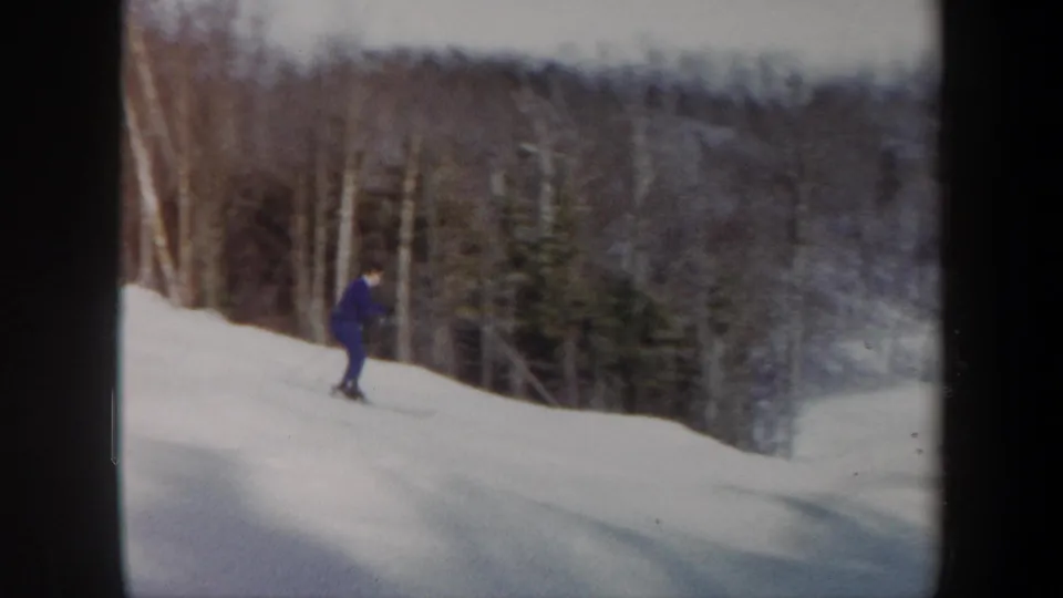 A blue-clad skier appears lower in frame on a snowy wooded slope, with film softness and muted color.
