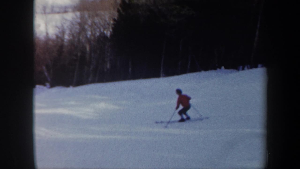 In Vermont 1962 home-movie footage, a skier in a red jacket crosses a snow-covered slope with dark woods behind.