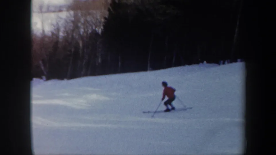 In Vermont 1962 home-movie footage, a skier in a red jacket crosses a snow-covered slope with dark woods behind.