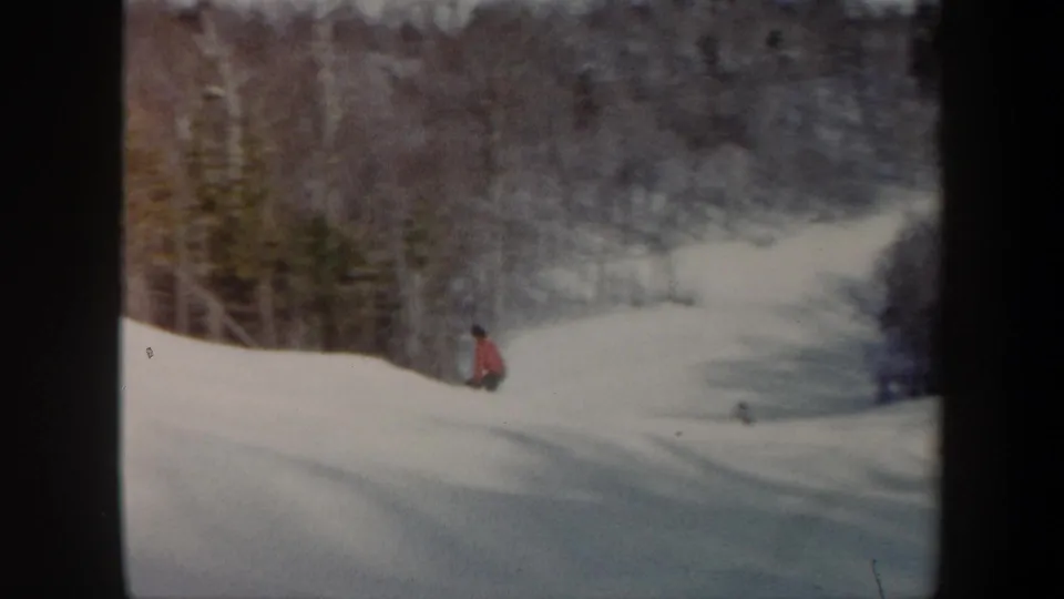 A distant skier in red is seen near the lower part of a snowy Vermont slope, with dark winter woods beyond.