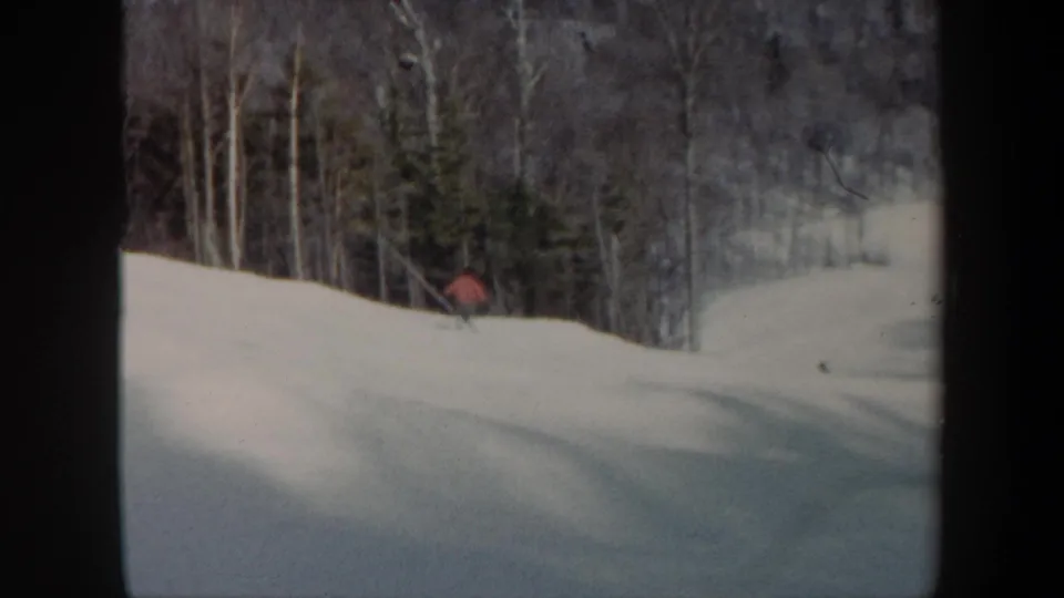 1962 Vermont 8mm frame showing a distant skier in red crossing a broad snow-covered ski slope with bare trees and evergreens behind.