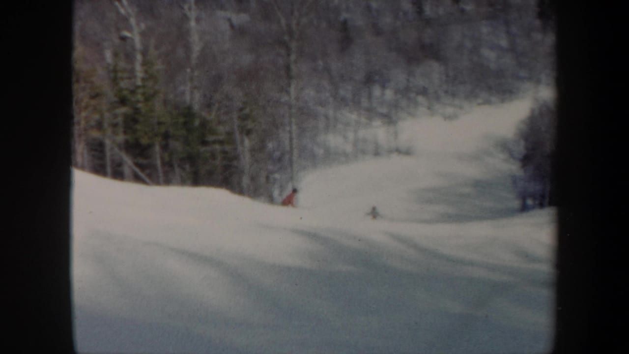 1962 Vermont ski slope seen in soft 8mm color, with distant skiers moving down open snow between wooded edges.