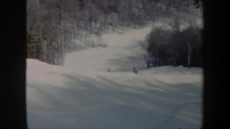 Wide 1962 Vermont home-movie view of a snow-covered ski run with a small red-clad skier and wooded hills behind.