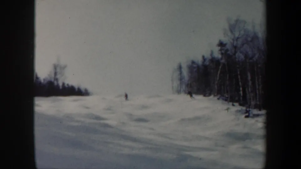 1962 Vermont ski footage frame with distant figures descending a white mogul slope beside a stand of bare trees.
