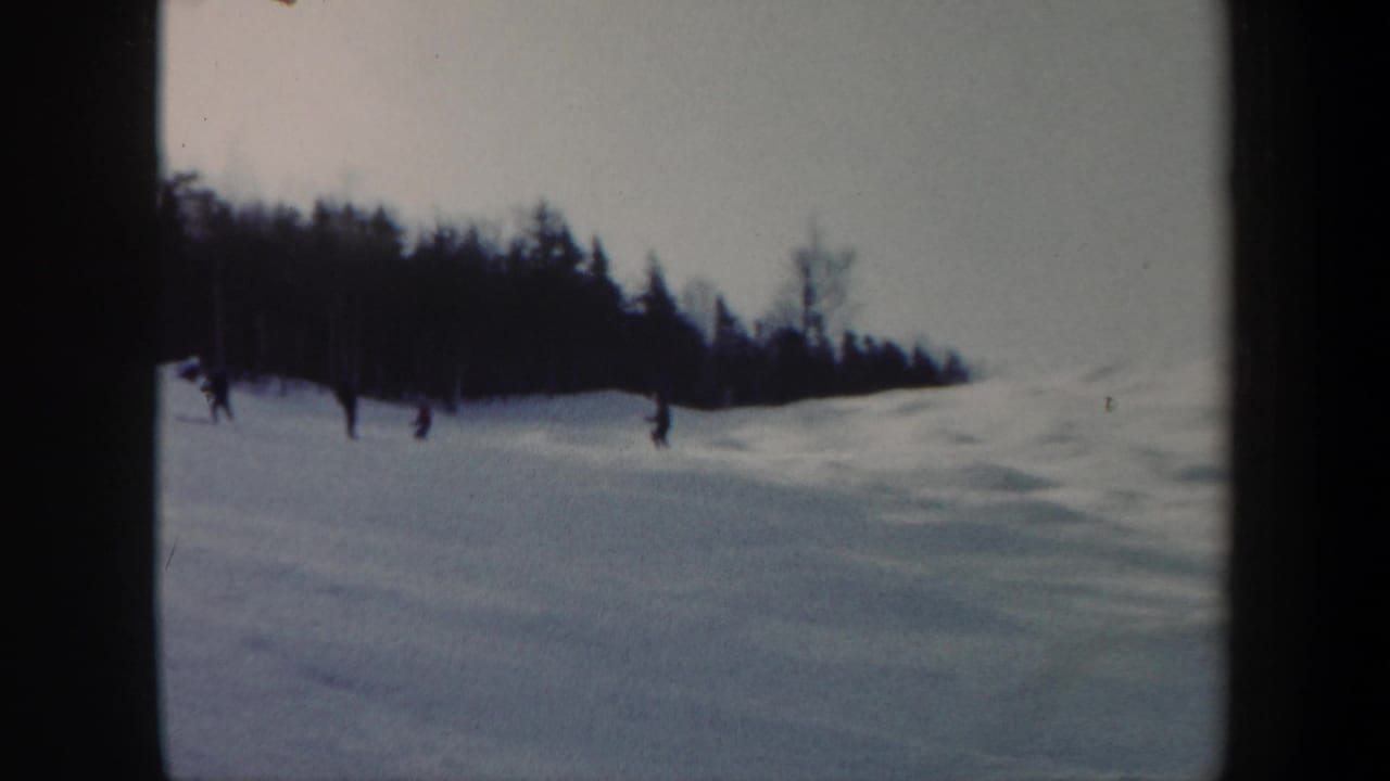 1962 Vermont home-movie frame showing distant skiers crossing a snowy slope near evergreen and leafless trees.