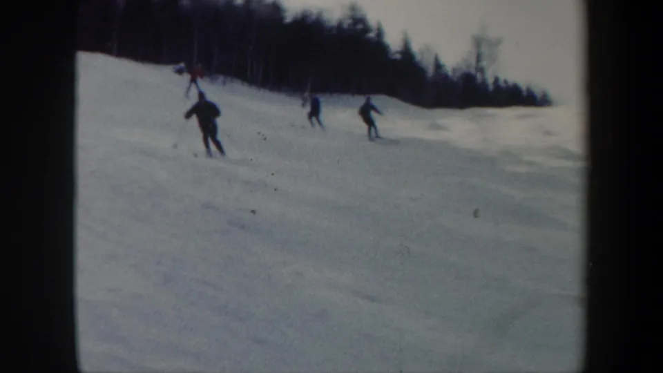 A closer skier and several others glide downhill across a snowy Vermont slope lined by winter trees.