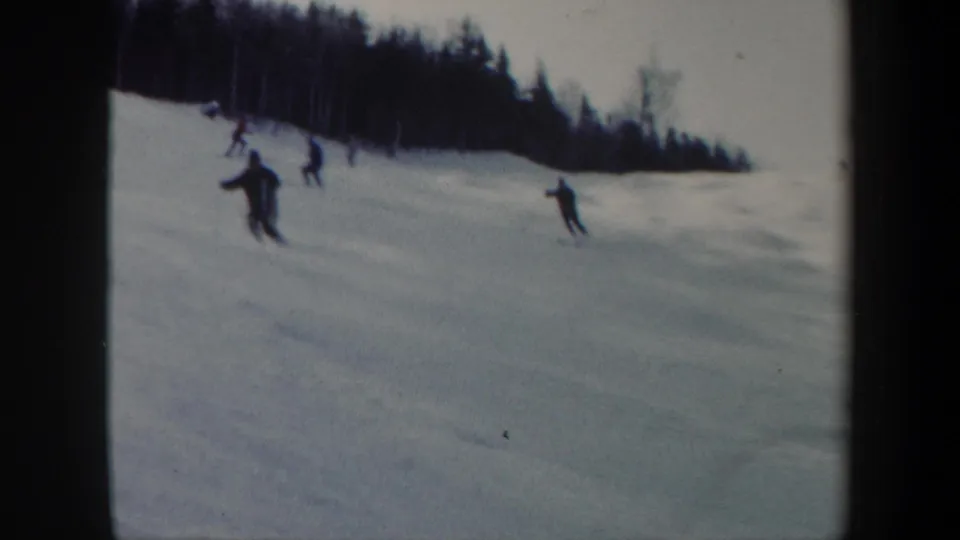 1962 Vermont 8mm frame showing several skiers in dark clothing moving downhill across a snowy slope near a forest edge.