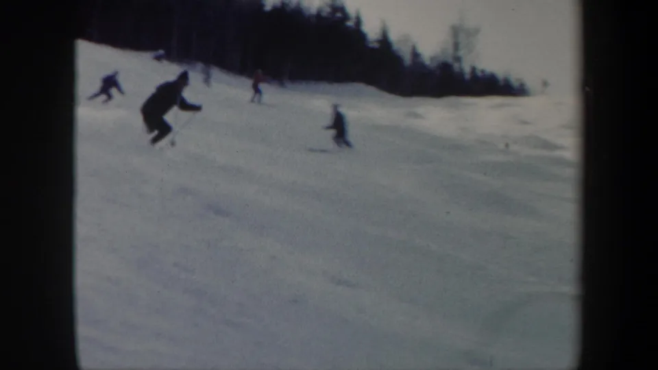 1962 Vermont home-movie frame with a foreground skier descending the slope and several smaller skiers near the treeline.