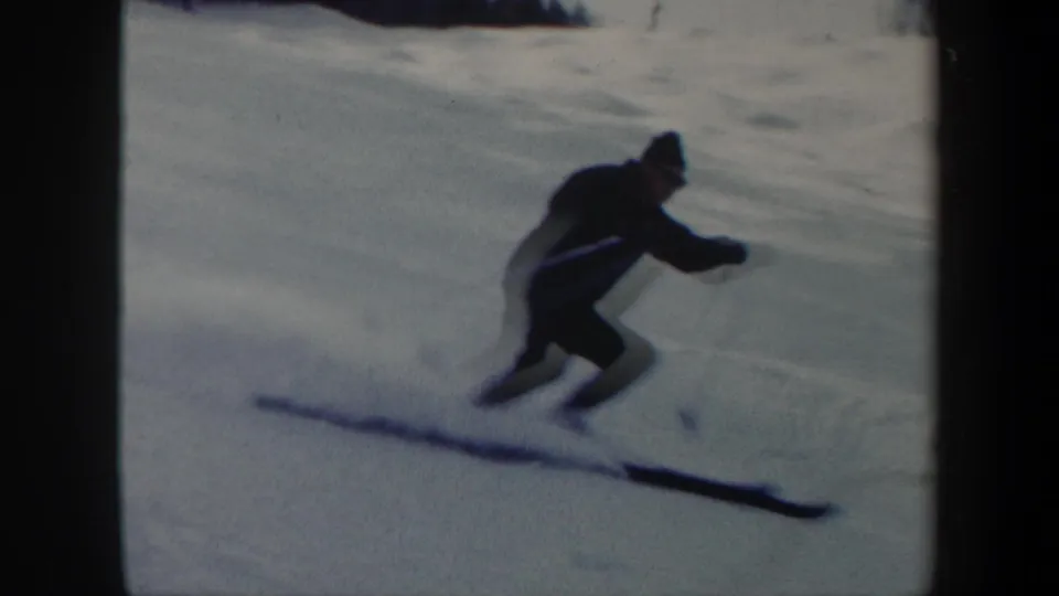 1962 Vermont home-movie frame with a skier close to the camera leaning into a downhill turn on long skis.