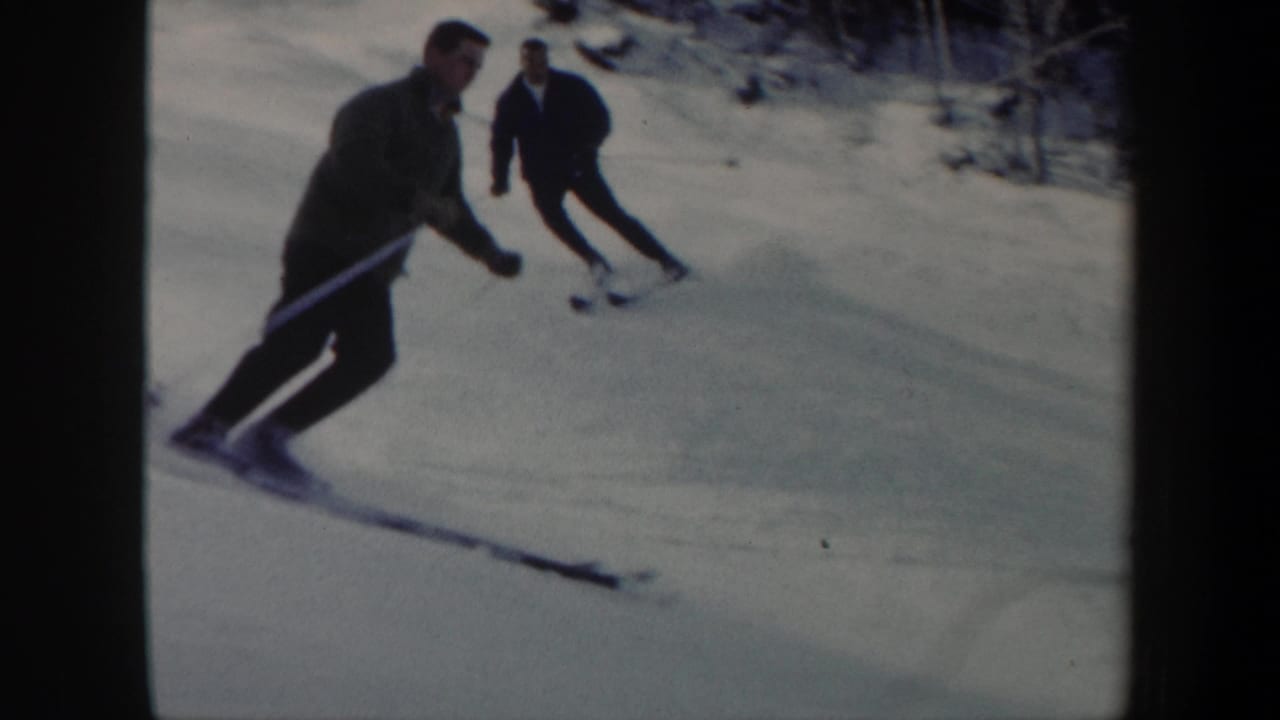 1962 Vermont 8mm frame showing two adult skiers moving downhill on a snowy slope bordered by bare trees.