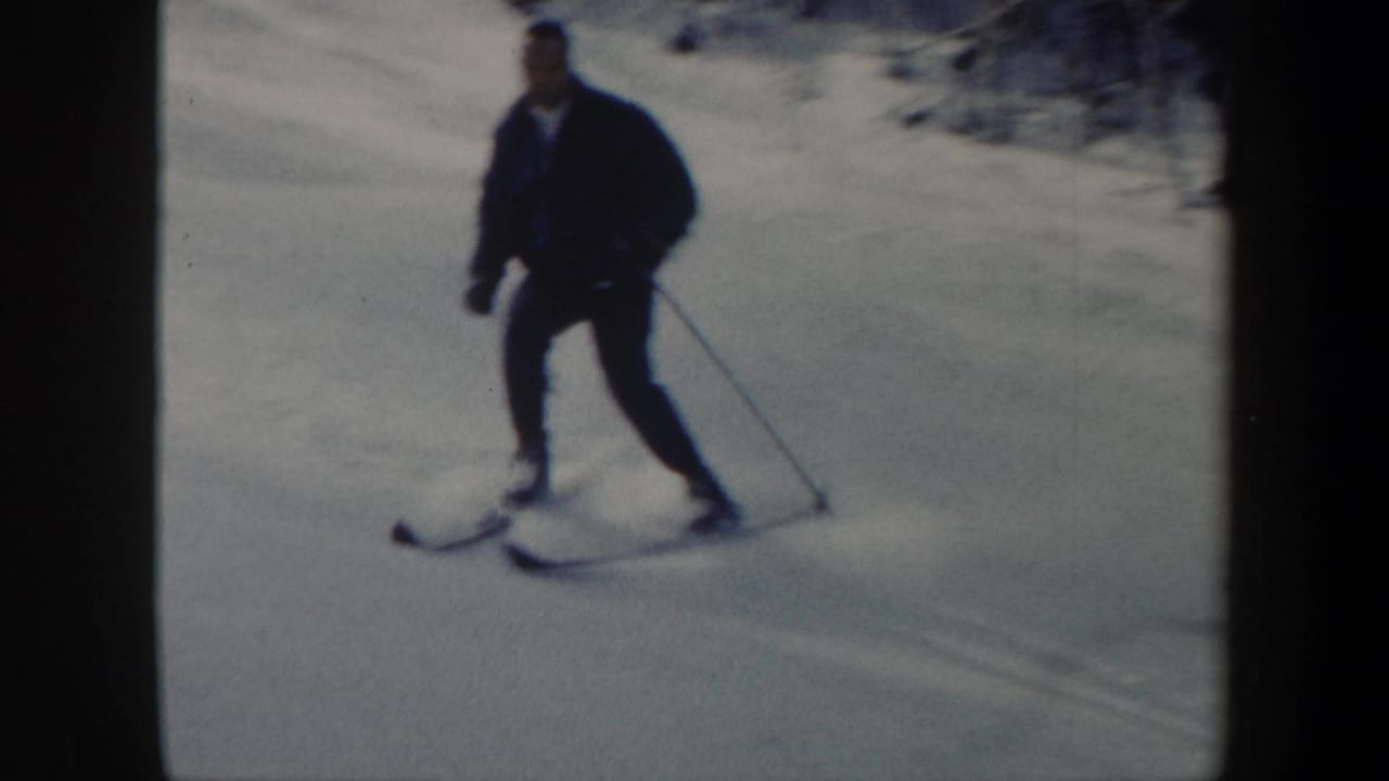 An adult skier in a dark jacket glides downhill with ski poles on a snowy slope.