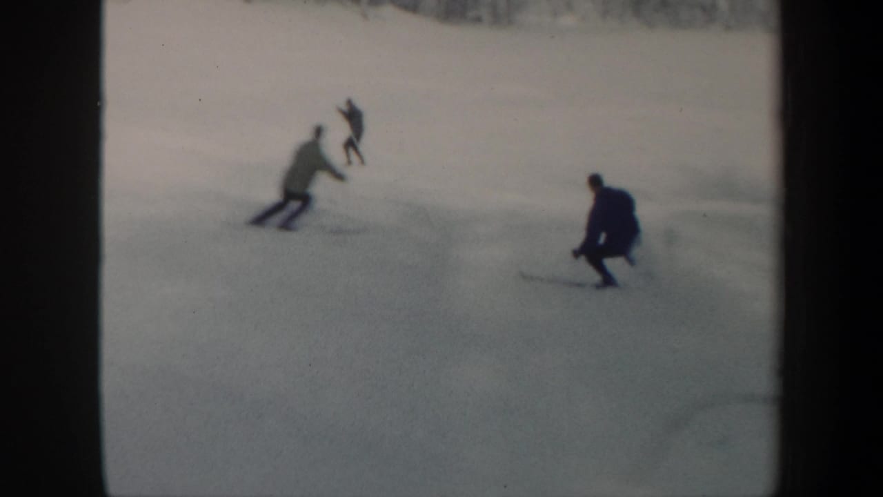 Distant skiers in 1962 Vermont move downhill across a pale snowfield with trees beyond the slope.