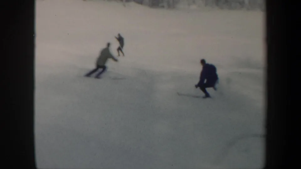 Distant skiers in 1962 Vermont move downhill across a pale snowfield with trees beyond the slope.
