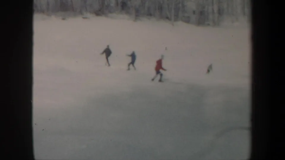 1962 Vermont home-movie frame showing a small group of skaters spread across a snowy frozen lake below bare trees.