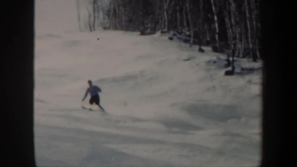 A distant skier descends a Vermont slope in 1962 home-movie footage, with trees and uneven snow visible behind.