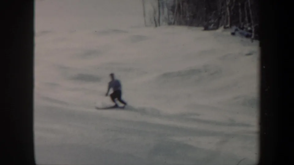 In 1962 Vermont home-movie footage, a distant adult skier moves across a snowy hillside near bare winter trees.