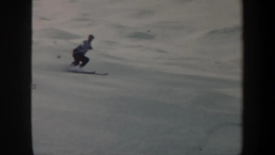 A skier in a light top and dark pants crouches into a turn on an open snowy Vermont slope in 1962 footage.