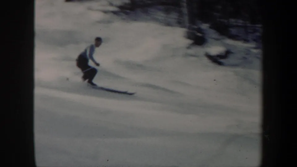 The adult skier appears closer in frame, leaning forward on long skis against a soft, grainy winter landscape.